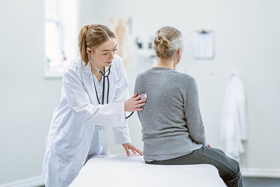 A female doctor holds her stethoscope to a patients chest as she listens to her lungs. She is wearing blue scrubs and has a white lab coat over top as she listens carefully.