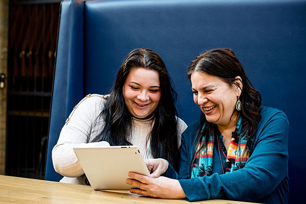 Seated side by side, an indigenous mother and daughter delve into a captivating video on their tablet, embracing the digital age together