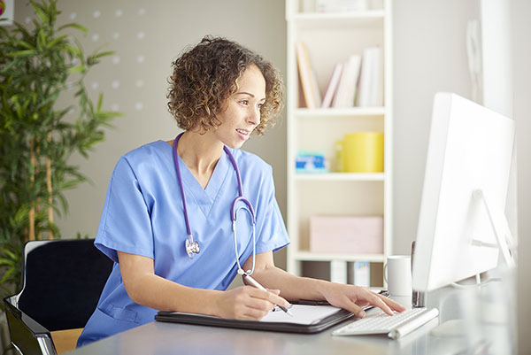 A female doctor sits at her desk and checks something on her computer She is wearing blue scrubs and stethoscope and seems happy in her work. She is lit by bright window light.