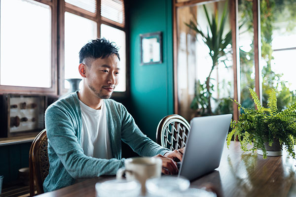 Smiling young Asian man sitting at dining room table at home, shopping online with laptop stock photo