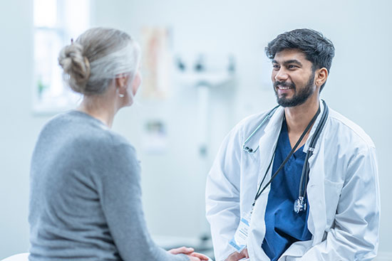 A senior woman sits up on an exam table as she meets with her doctor to discuss her health concerns. Her male doctor of Middle Eastern decent is seated across from her in a white lab coat as the two talk.