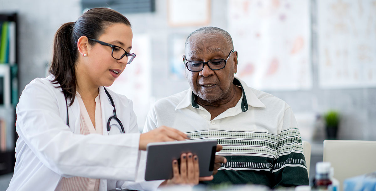 A senior man of African descent is indoors in a hospital room. He is watching his female doctor using a tablet computer. She is explaining a medication schedule to him.