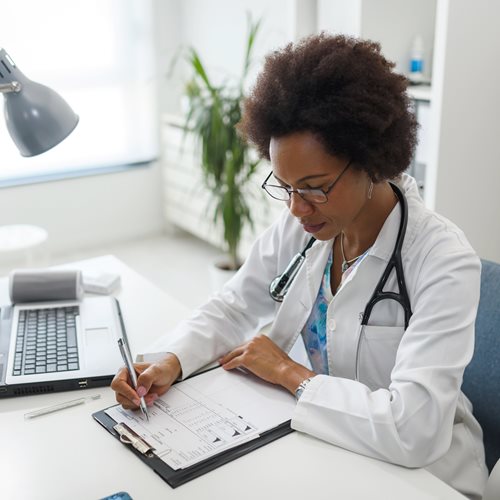 photo of a female physician, sitting at a desk, writing on a clipboard chart